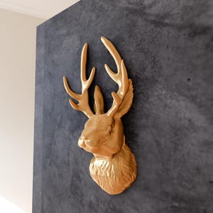 May include: A gold-coloured deer head wall mount with antlers. The deer head is mounted on a dark grey textured wall.