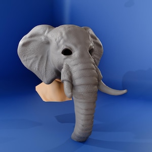 Può includere: Una maschera da elefante grigia con grandi orecchie, una lunga proboscide e zanne. La maschera è posta su uno sfondo blu ed è attaccata a una base color beige.