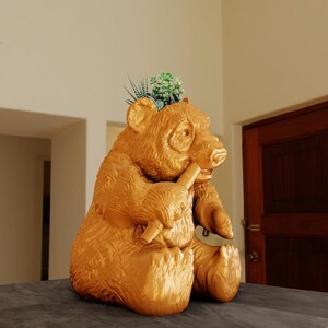 Könnte beinhalten: Ein goldfarbener Panda-Pflanztopf mit einer kleinen Sukkulentenpflanze, die aus der Oberseite wächst. Der Topf steht auf einer grauen Oberfläche.