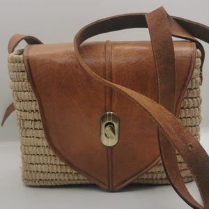 Puede incluir: Bolso bandolera marrón y beige. El bolso tiene un cuerpo de paja tejida con una solapa y correa de cuero marrón. La solapa tiene una forma decorativa y un cierre dorado. La correa es ajustable.
