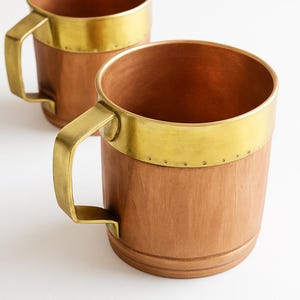 Può includere: Due tazze di legno con bordi e manici in ottone. Le tazze hanno una finitura in legno marrone chiaro e una fascia in ottone color oro sulla parte superiore. Anche i manici sono in ottone. Le tazze sono su uno sfondo bianco.