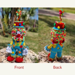 May include: A crocheted clown doll with a colorful hat and a red nose. The doll is wearing a blue and yellow outfit with a white collar. The doll is standing on a rock.