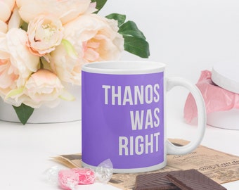 Könnte beinhalten: Weiße Keramiktasse mit einem lila Band mit dem weißen Text "THANOS WAS RIGHT". Die Tasse ist von Blumen, Süßigkeiten und Schokolade umgeben.