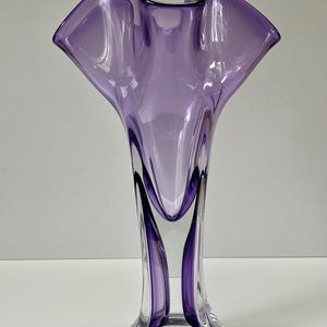 Op de afbeelding: Een hoge, elegante vaas met een uitlopende, gerimpelde bovenkant in een doorschijnende lavendelkleur. De vaas heeft een heldere, afgeronde basis en een slanke, langwerpige body. Het ontwerp heeft vloeiende, organische lijnen, waardoor een gevoel van beweging en verfijning ontstaat. De vaas is ongeveer 30 cm hoog.