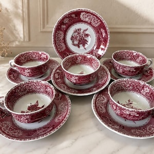 Può includere: Un set di cinque tazze da tè e piattini in ceramica con un motivo floreale rosso scuro e bianco. Ogni tazza è dotata di un piattino abbinato con dettagli botanici intricati e bordi decorati, disposti in una composizione a livelli.