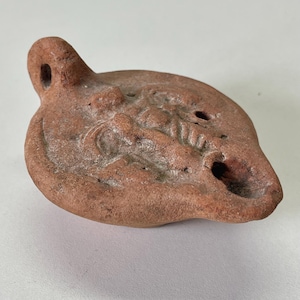 Puede incluir: Lámpara de aceite de terracota marrón rojizo con un relieve detallado. La lámpara ovalada tiene un asa pequeña y un pico. La superficie rugosa sugiere un objeto antiguo o vintage. Probablemente de importancia histórica.