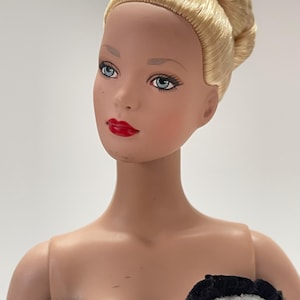 Op de afbeelding: Een pop met blond haar in een knot. De pop heeft blauwe ogen, rode lippen en een lichte teint. De pop draagt een lichtblauwe jurk met een zwarte gefranjerde rand.
