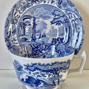 Può includere: Un set di tazza e piattino in porcellana blu e bianca. La tazza e il piattino presentano un design paesaggistico con alberi, edifici e un ponte. Il set è esposto su un supporto rotondo trasparente. Il design è in stile classico.