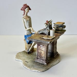 Può includere: Una statuetta dettagliata raffigura una donna seduta vicino a una scrivania. La donna indossa un top bianco e una gonna blu. La scrivania ha una pila di carte, una tazzina e un oggetto decorativo. La base è di colore beige chiaro.