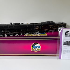 Może przedstawiać: Czarna miniaturowa lokomotywa parowa z numerem 2124 i napisem READING na tenderze. Stoi na fioletowym pudełku z logo MTH Electric Trains. Dołączona jest instrukcja obsługi lokomotywy parowej Premier T-1 Reading.