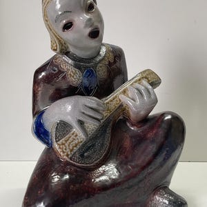 May include: A ceramic figurine of a person playing a guitar. The figurine is painted in shades of brown, blue, and white. The person is wearing a long, flowing robe and has a cheerful expression on their face.