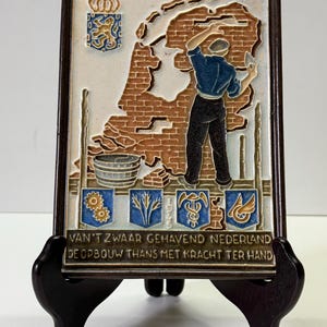 May include: A rectangular ceramic wall plaque in a dark brown frame featuring a relief of a person building a brick wall, the Dutch coat of arms, and four blue shields. The text reads: VAN'T ZWAAR GEHAVEND NEDERLAND DE OPBOUW THANS MET KRACHT TER HAND.