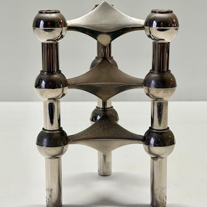 May include: A silver-toned, three-legged candle holder with a geometric design. The candle holder features spherical accents and triangular elements, creating a modern, minimalist aesthetic. The piece is made of metal and has a polished finish.
