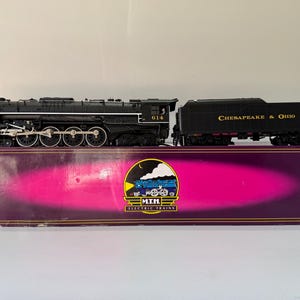 Può includere: Una locomotiva a vapore in miniatura M.T.H. Electric Trains nera con tender, recante il numero 614 sulla cabina. Il tender riporta la scritta Chesapeake & Ohio in lettere dorate. Il treno è posizionato su una scatola viola con un logo.