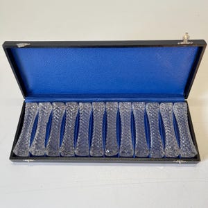May include: A set of 16 clear glass butter knives in a black box with a blue velvet lining. The knives have a diamond pattern.