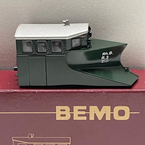 Peut inclure: Une modèle de train de déneigement vert et gris avec le texte "Rh.B. R.3" sur le côté. Le train est fabriqué par BEMO.