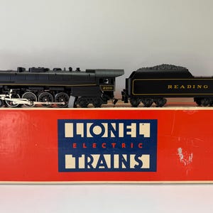 Puede incluir: Una locomotora de vapor eléctrica negra Lionel con ténder, letras doradas que dicen READING y el número 2100. El tren está colocado sobre una caja rectangular roja con una etiqueta azul y blanca que dice LIONEL ELECTRIC TRAINS.