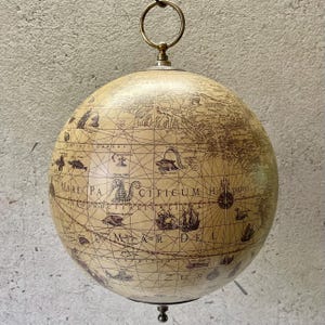 Könnte beinhalten: Ein Globus im Vintage-Stil mit cremefarbener Oberfläche und detaillierter brauner Kartografie. Der Globus ist an einem Messingring aufgehängt und zeigt Illustrationen von Schiffen und Meereskreaturen. Ein dekoratives Element für Zuhause oder Büro.