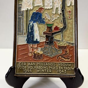 May include: A rectangular ceramic wall plaque featuring a relief scene of a person cooking on a stove with laundry hanging above. The text reads: EER AAN HOLLANDS VROUW VOOR VOLHARDING MOED EN TROUW 1944 WINTER 1945. The plaque has a dark brown frame and a multi-coloured finish.