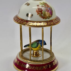 Op de afbeelding: Een porseleinen vogelkooi met een gouden rand aan de basis en een koepelvormige bovenkant. De kooi is versierd met een bloemenpatroon en een scène van twee mensen in een tuin. Een kleine blauwe en gele vogel zit in de kooi.