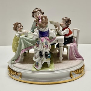 May include: A porcelain figurine depicting four children sitting around a table, with a green tablecloth and a pink tablecloth on the chair. The children are dressed in colorful clothing and are holding plates and cups. The figurine is on a round base with gold accents.