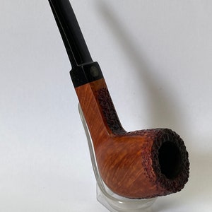 Peut inclure: Une pipe en bois brun avec un embout noir et une finition texturée. La pipe est posée sur un support en plastique transparent.