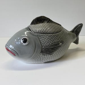 Può includere: Un piatto da portata a forma di pesce in ceramica grigia, con pinna dorsale e coda grigio scuro. Il piatto ha un coperchio ed è rifinito con squame, bocca rossa e occhi neri. Adatto per servire cibo.