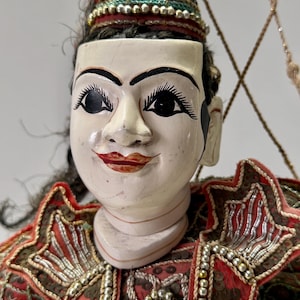 Op de afbeelding: Een close-up van een traditionele Aziatische pop. De pop heeft een geschilderd gezicht met zwarte ogen, wenkbrauwen en een kleine rode mond. Ze draagt een decoratieve hoofdtooi en een rood en goud geborduurd kostuum. Het haar van de pop is zwart en krullend.