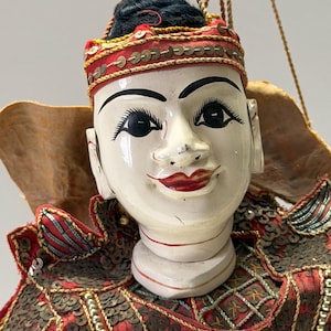 Op de afbeelding: Een traditionele Aziatische pop met een geschilderd wit gezicht, zwart haar en rode lippen. De pop draagt een rode en gouden hoofdtooi en een gedetailleerd rood en goud kostuum. De pop hangt aan touwtjes.