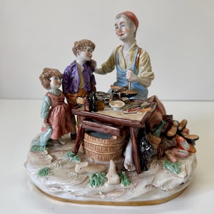 Puede incluir: Figurilla de porcelana que representa a un zapatero con dos niños. El zapatero, con delantal azul y gorro rojo, trabaja en una mesa con herramientas. Los niños visten ropa colorida. La base es un paisaje detallado.
