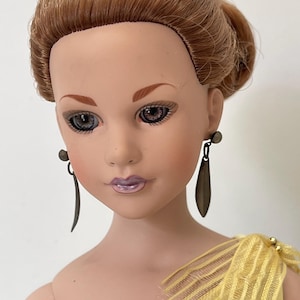 May include: A vintage doll with auburn hair in an updo. The doll has painted facial features, including eyes and lips. She wears dark metal drop earrings and a yellow dress with a decorative shoulder detail.