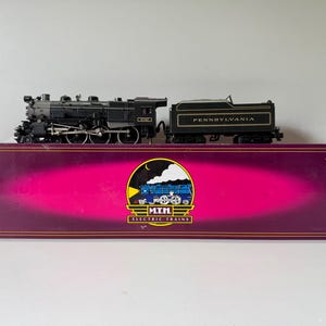 Pode incluir: Uma locomotiva a vapor em miniatura preta e cinzenta com um tender rotulado Pennsylvania, pousada sobre uma caixa retangular roxa comprida. A caixa apresenta um logótipo circular central com um gráfico de comboio azul e o texto M.T.H. ELECTRIC TRAINS.