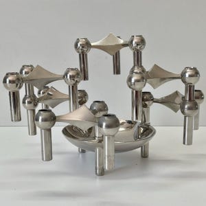 May include: A silver-toned metal candle holder with multiple candle cups and geometric shapes. The design features a central bowl and several elevated candle holders, creating a modern, artistic display. The piece is made of shiny metal.