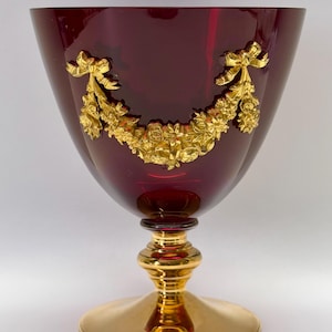Puede incluir: Una copa de cristal rojo rubí con base y tallo dorados. La copa está adornada con una guirnalda floral dorada y un diseño de lazo. La copa tiene un cuenco ancho y una base redonda.