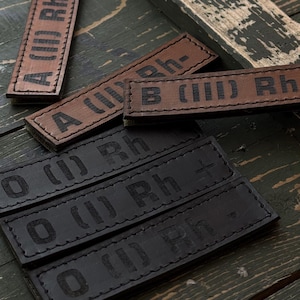 Puede incluir: Parches de cuero en marrón y negro con tipos de sangre. Los parches muestran tipos de sangre como A (II) Rh+, B (III) Rh-, y O (I) Rh+. Los parches rectangulares tienen bordes cosidos y texto en relieve.