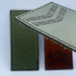 May include: Three rectangular leather patches with stitched edges. One light gray patch is angled in the foreground, featuring a debossed chevron pattern. Two additional patches, one dark green and one reddish-brown, lie flat in the background.