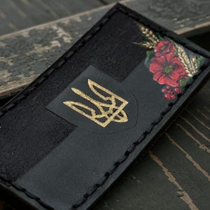 Peut inclure: Portefeuille en cuir noir avec un emblème doré et des motifs floraux. Le portefeuille a une bordure cousue et une forme rectangulaire. L'œuvre d'art comprend des fleurs rouges et du blé. L'emblème est un trident doré.