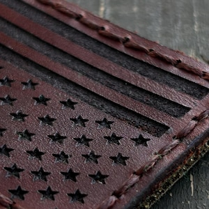 May include: A close-up of a leather patch depicting the American flag. The patch has a distressed appearance with a dark red background and black stars and stripes. The edges are stitched with a contrasting thread.
