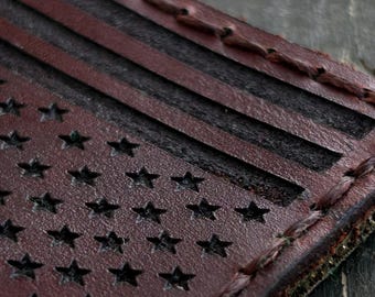 Könnte beinhalten: Nahaufnahme eines Lederaufnähers mit dem Design der amerikanischen Flagge. Der Aufnäher hat einen Used-Look mit dunkelrotem Hintergrund und schwarzen Sternen und Streifen. Die Kanten sind mit einem Kontrastfaden genäht.