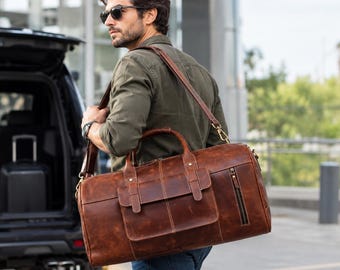 May include: A brown leather duffel bag featuring a large front flap pocket with two buckle-style closures, a vertical zippered side pocket, two top handles, and a detachable shoulder strap with brass-toned metal hardware clips.