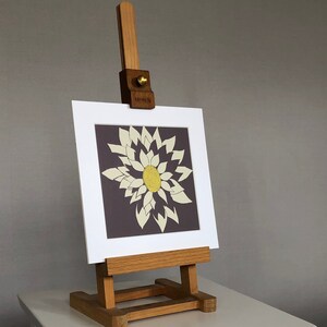 May include: A small wooden artist's easel holding a framed artwork. The artwork features a stylized flower with cream-colored petals and a yellow center against a dark background. The easel is made of light-colored wood and sits on a white surface.