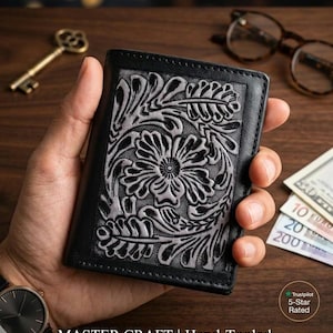 Könnte beinhalten: Schwarzes Lederportemonnaie mit handgearbeitetem Blumen- und Farnmuster. Das Portemonnaie hat ein detailliertes, geprägtes Silbermuster auf schwarzem Hintergrund. Der Text "MASTER CRAFT | Hand-Tooled Floral & Fern Leather Wallet" ist sichtbar.