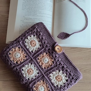 May include: A handmade crochet book cover in a deep purple hue, featuring a floral pattern with white and peach-colored accents. A matching crochet bookmark with a flower and leaf design rests in an open book. A wooden button secures the cover.