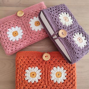 Può includere: Tre copertine per libri all'uncinetto con motivi a margherita. Le copertine sono in tonalità di rosa, viola e arancio. Ogni copertina ha un bottone di legno.