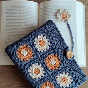 Può includere: Un astuccio blu all'uncinetto con un motivo a margherita nei colori arancione, bianco e crema. L'astuccio ha una chiusura con bottone in legno e un segnalibro a margherita abbinato. L'astuccio è aperto su un libro.