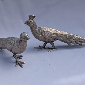 May include: Two silver metal bird ornaments. One is a peacock with a long, detailed tail and a crown-like crest. The other is a smaller bird with a shorter tail. Both have detailed feather textures and stand on three-toed feet.