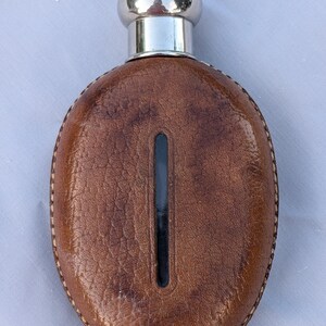 Pode incluir: Uma petisqueira de formato oval envolta em couro castanho texturizado com uma janela de visualização vertical preta no centro. A petisqueira apresenta uma tampa e gargalo de metal prateado polido, com costuras visíveis na borda externa.