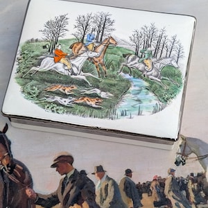 Puede incluir: Caja de baratijas rectangular blanca con una ilustración de una escena de caza. La caja presenta jinetes a caballo, perros y un arroyo. La ilustración es en tonos verdes, marrones y azules. La caja tiene un borde plateado.