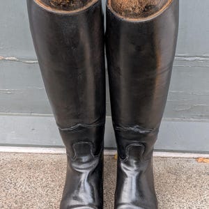 以下が含まれることがあります： 木製のシューツリーが挿入された、背の高い黒いレザー製の乗馬ブーツ一足。各ブーツは滑らかで光沢のある仕上げ、構造化されたシャフト、そして木製インサートの上部に取り付けられた真鍮色の金属リングが特徴です。