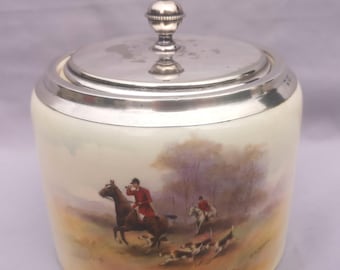 Puede incluir: Tarro de cerámica color crema con tapa y pomo plateados. El tarro presenta una escena pintada a mano de caza del zorro con jinetes a caballo, perros y un fondo de paisaje. La tapa y el borde son plateados.